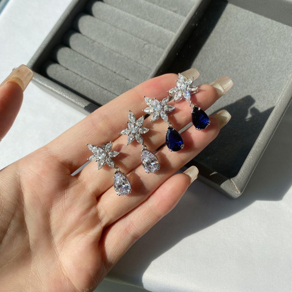 Two pairs of silver drop earrings featuring pear-shaped clear and blue stones with intricate floral designs above the earrings.