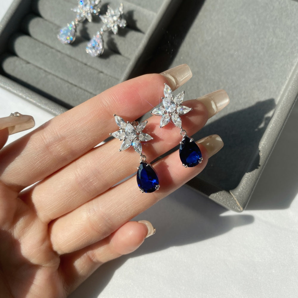 A pair of silver drop earrings featuring pear-shaped blue stones with intricate floral designs above the earrings.