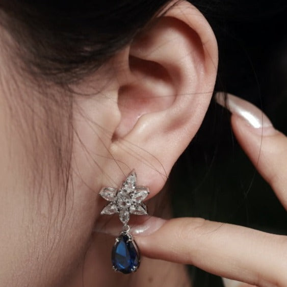 A silver drop earring featuring pear-shaped blue stone with intricate floral designs above the earring.