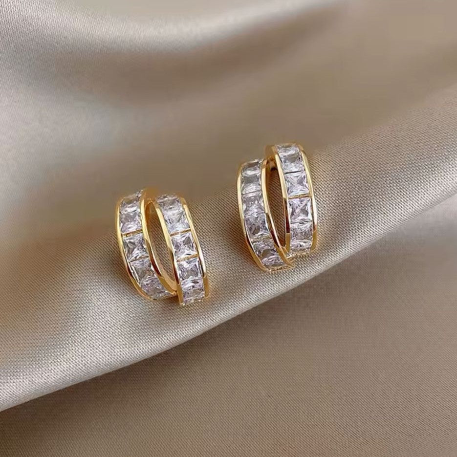 A pair of gold-tone huggie hoop earrings with a double row of square-cut crystals, displayed on a satin surface.