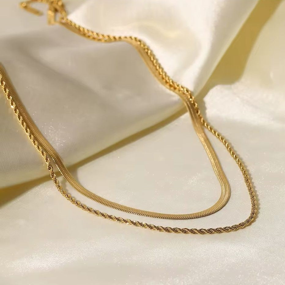 A gold colored twin chain necklace displayed over white fabric in the background.