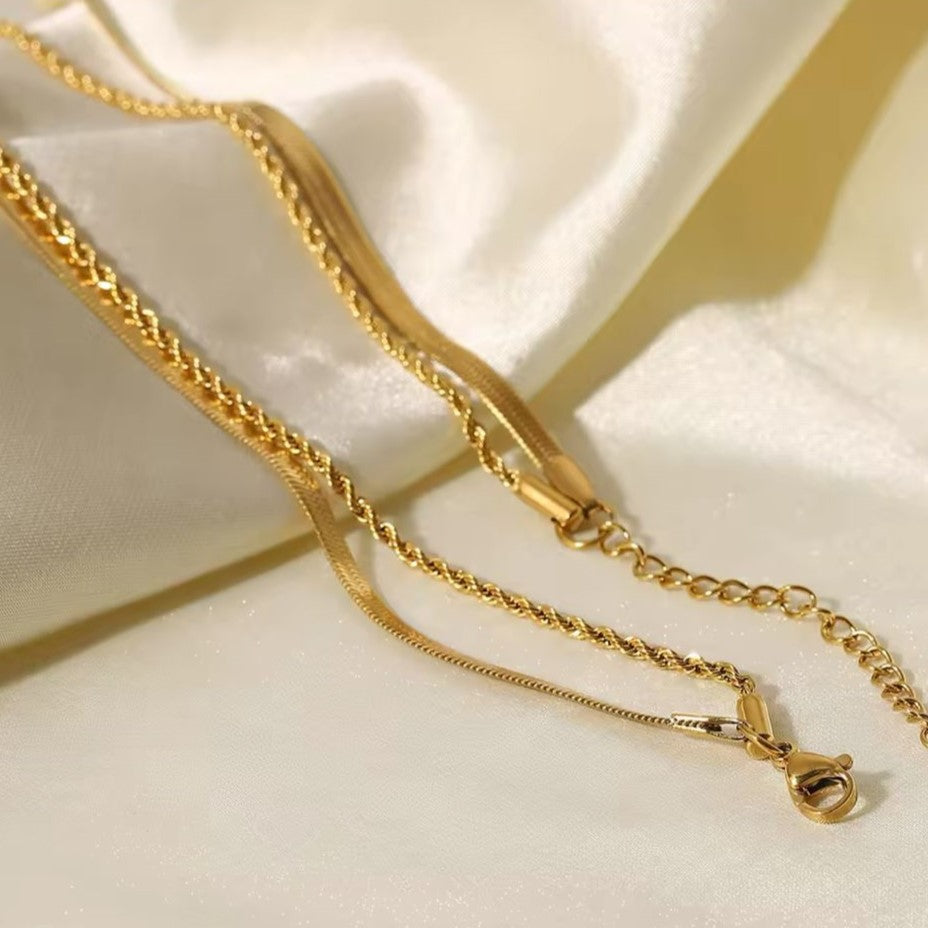 A gold colored twin chain necklace displayed over white fabric in the background.