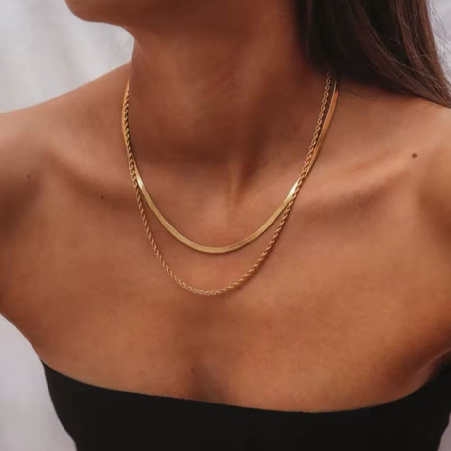 A gold colored twin chain necklace displayed over a women's neck with blurred background.