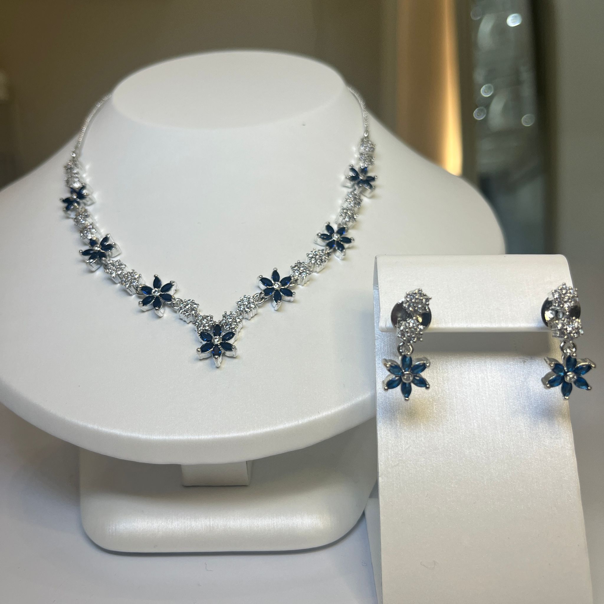 A jewelry set featuring a floral-inspired necklace with blue and white stones and matching drop earrings, displayed on a white mannequin.