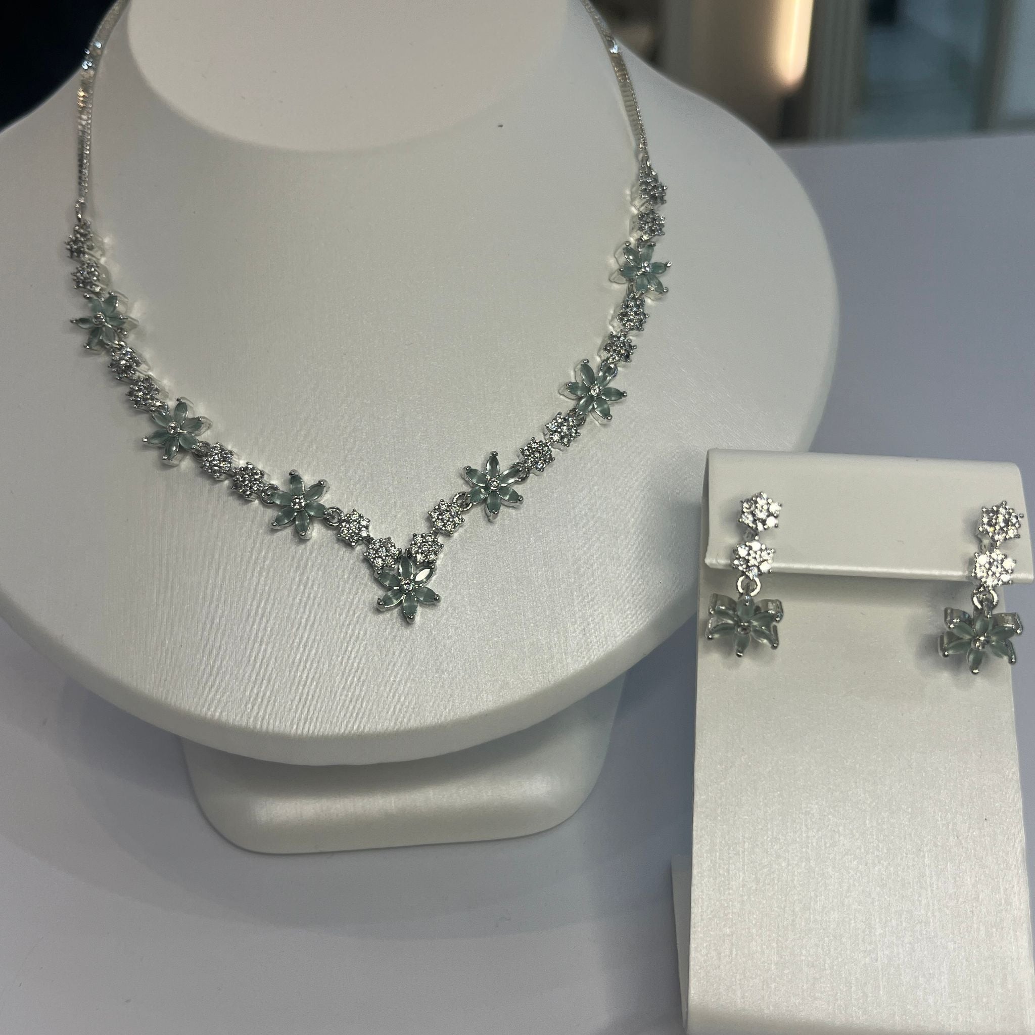A jewelry set featuring a floral-inspired necklace with green and white stones and matching drop earrings, displayed on a white mannequin.