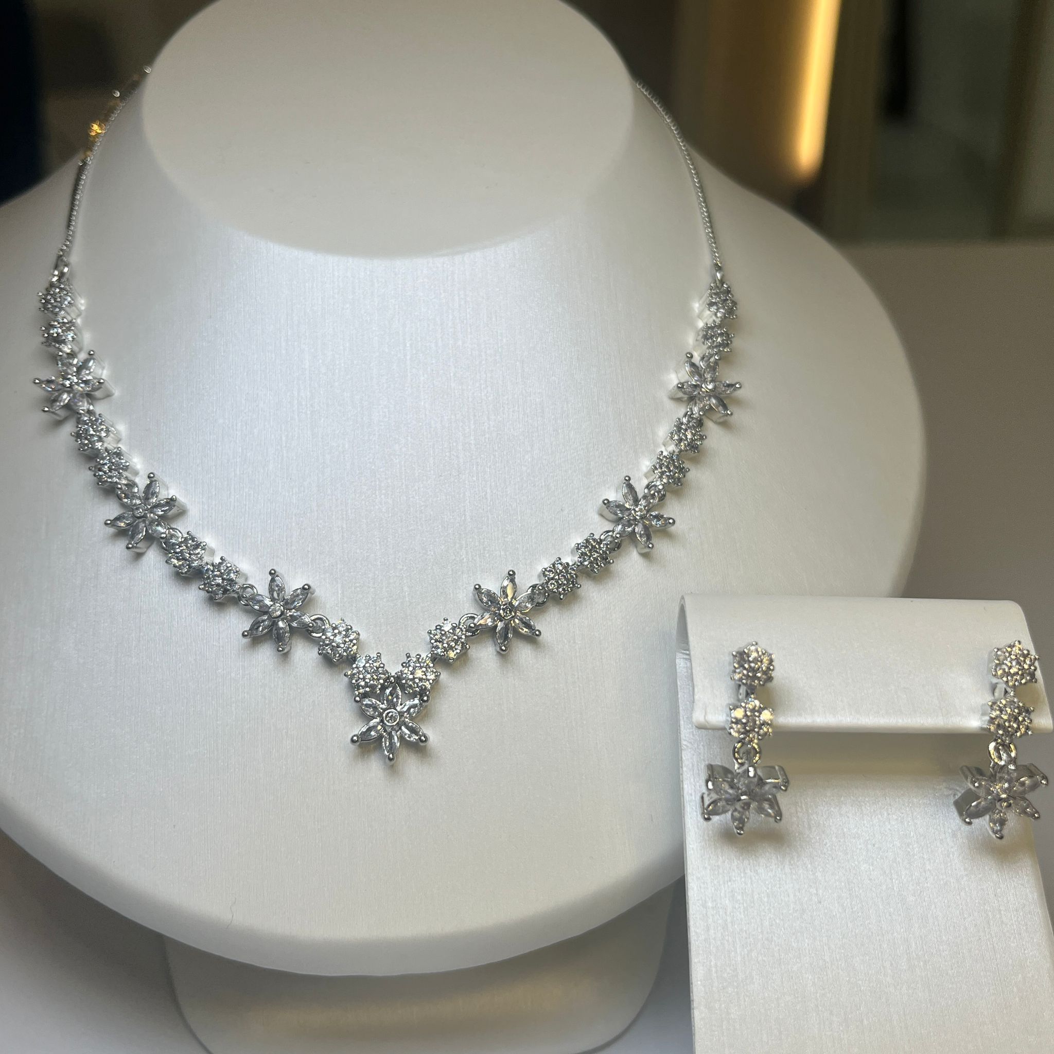 A jewelry set featuring a floral-inspired necklace with clear white stones and matching drop earrings, displayed on a white mannequin.