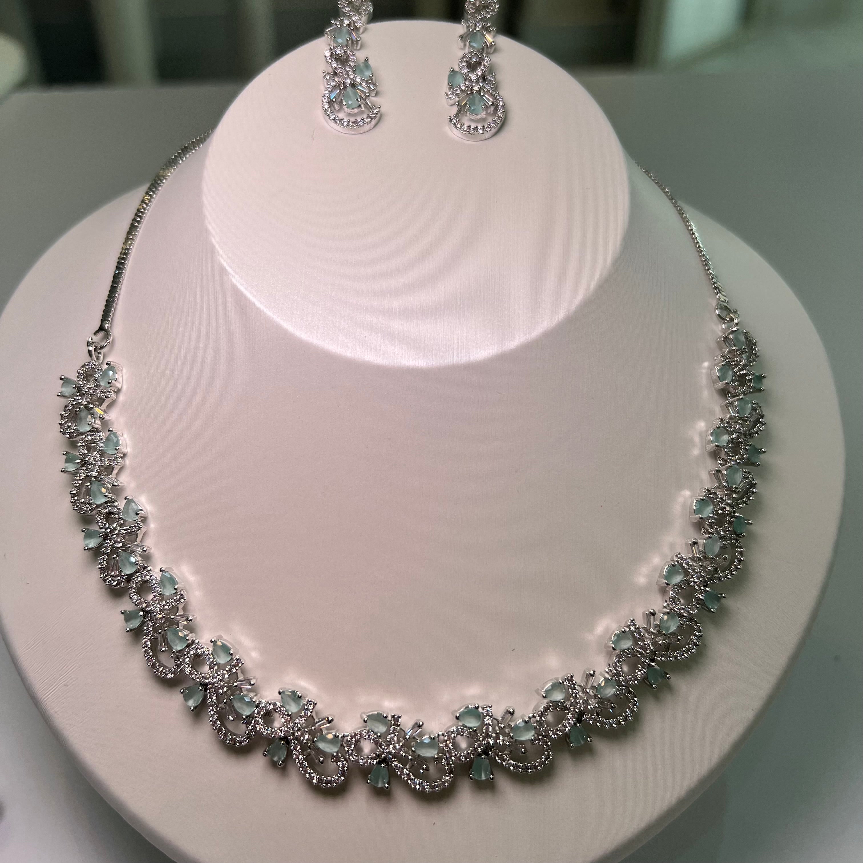 A necklace and earrings set displayed on a mannequin, featuring silver-tone chains with floral-inspired patterns and green-colored stones.