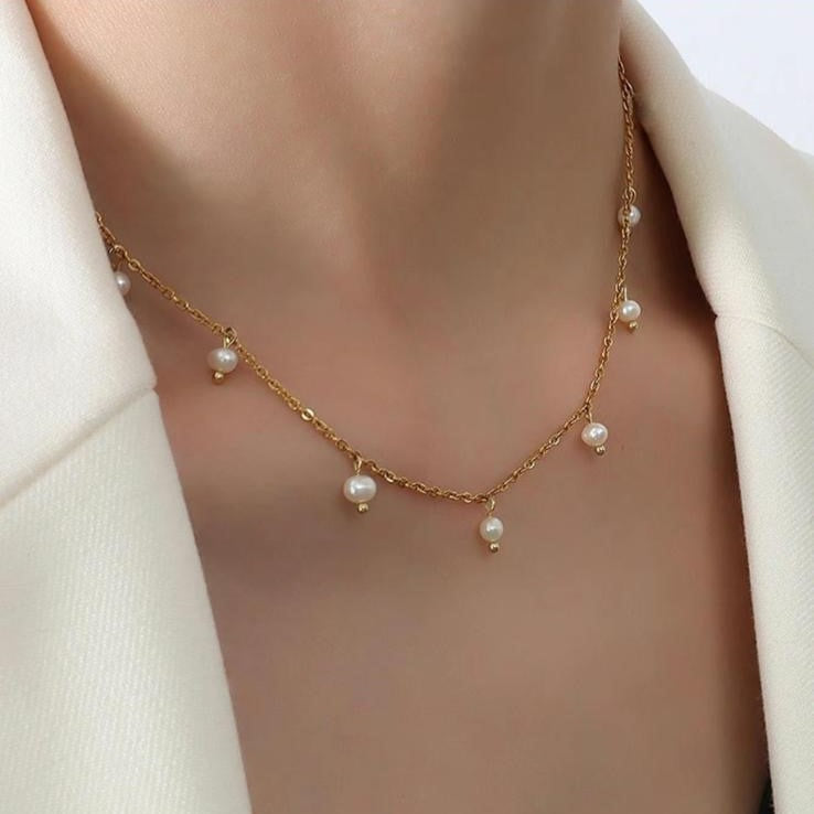 A gold-colored stainless steel necklace with pearl-like beads, displayed on a women's neck.
