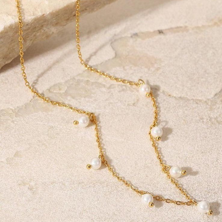 A gold-colored stainless steel necklace with faux pearl drop pendants on a beige background.