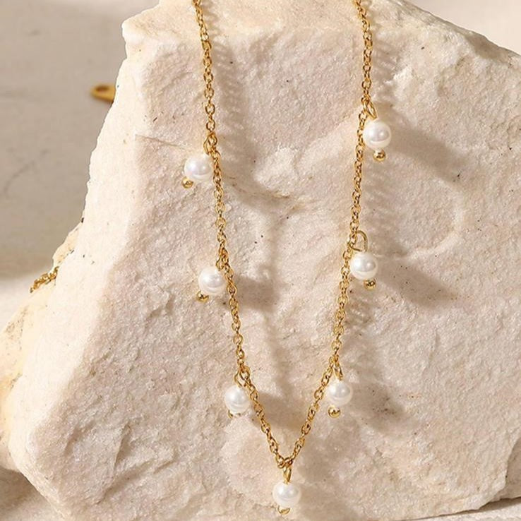 A gold-colored stainless steel necklace with faux pearl drop pendants on a beige background.