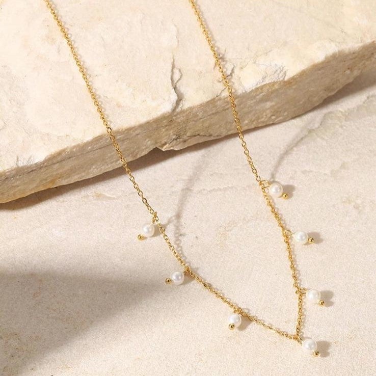 A gold-colored stainless steel necklace with faux pearl drop pendants on a beige background.