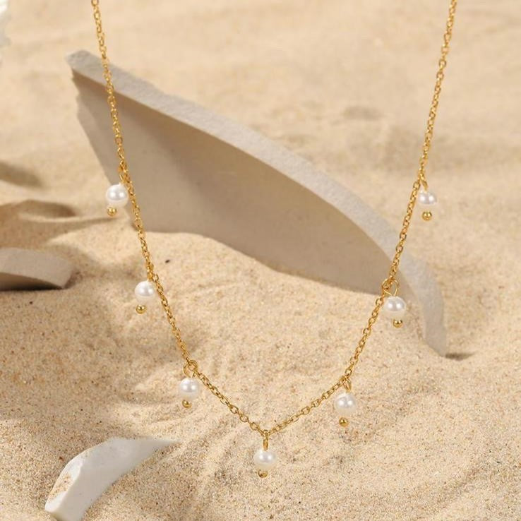 A gold-colored stainless steel necklace with faux pearl drop pendants on a beige background.