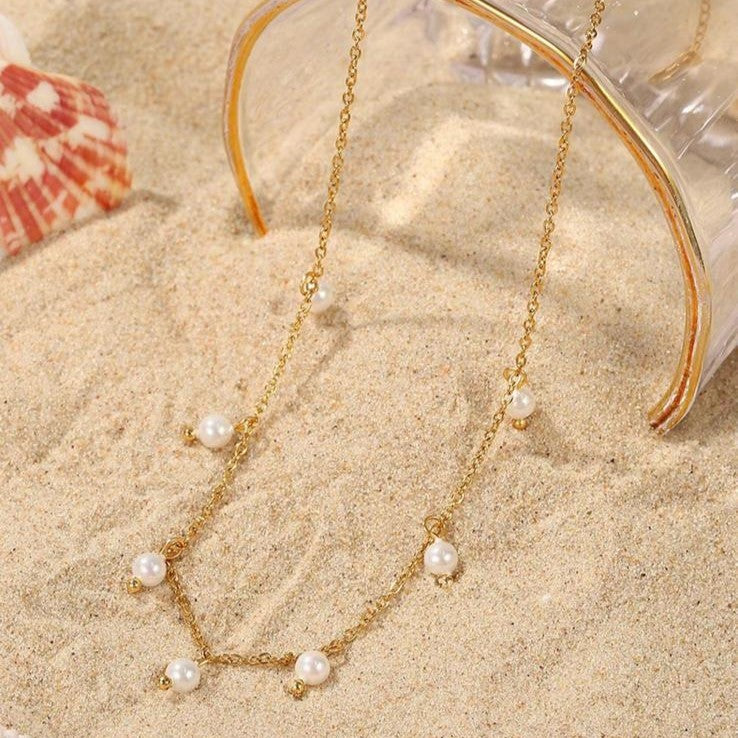 A gold-colored stainless steel necklace with faux pearl drop pendants on sand in the background.