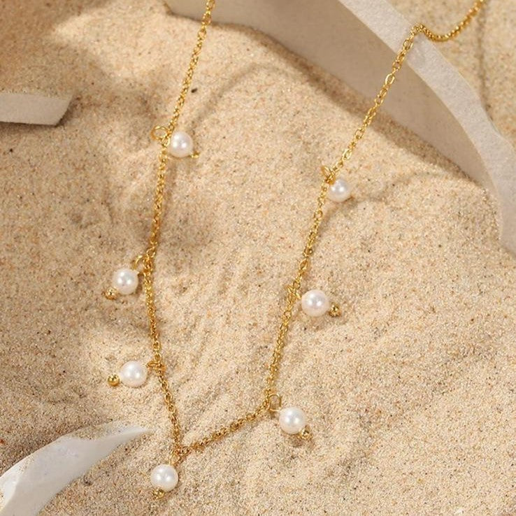 A gold-colored stainless steel necklace with faux pearl drop pendants on sand in the background.