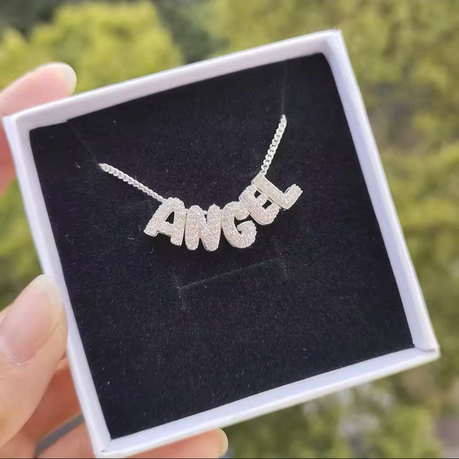 A personalized bubble letter charm necklace in silver tone, with necklace displaying charms, displayed on a box.