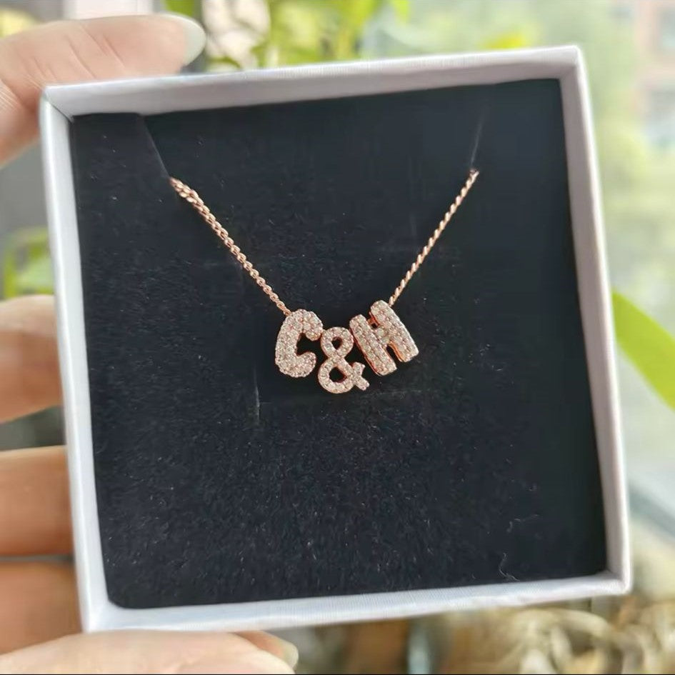 A personalized bubble letter charm necklace in gold tone, with necklace displaying charms, displayed on a box.