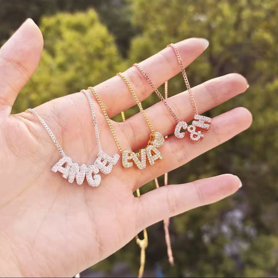 Three personalized bubble letter charm necklaces in bronze, silver and gold tone, with each necklace displaying charms in different sizes and styles, set against a blurred background on a hand.