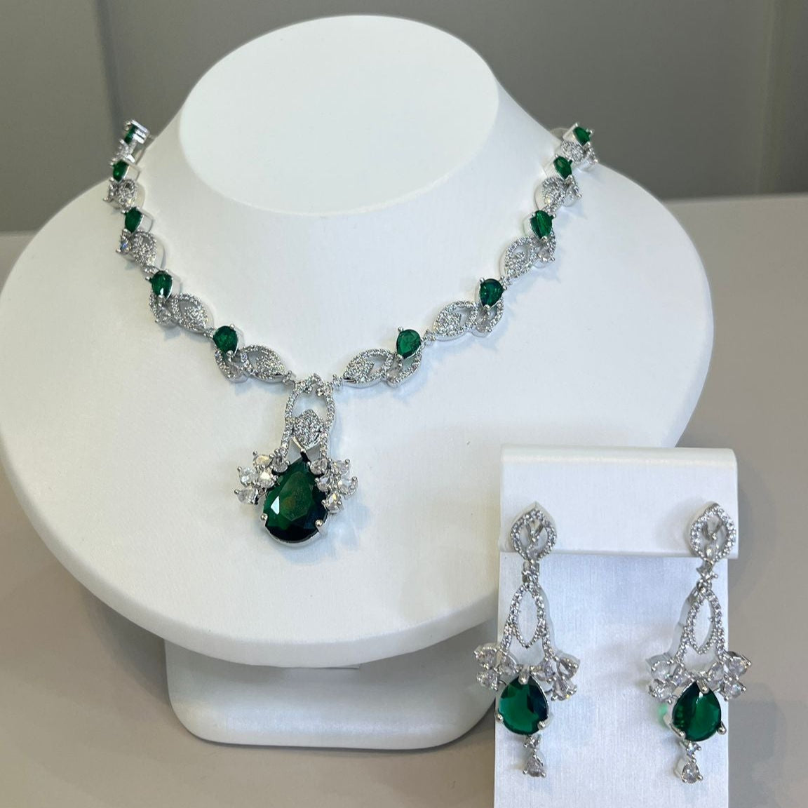 A luxurious silver necklace with a green gemstone centerpiece and sparkling crystals, accompanied by matching earrings, displayed on a white bust.