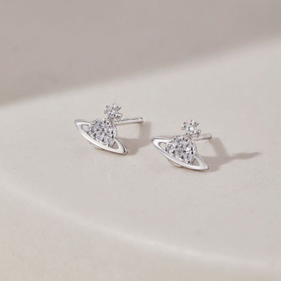 A pair of silver-colored stud earrings featuring a cosmic orbit design with sparkling stones, displayed against a light background.