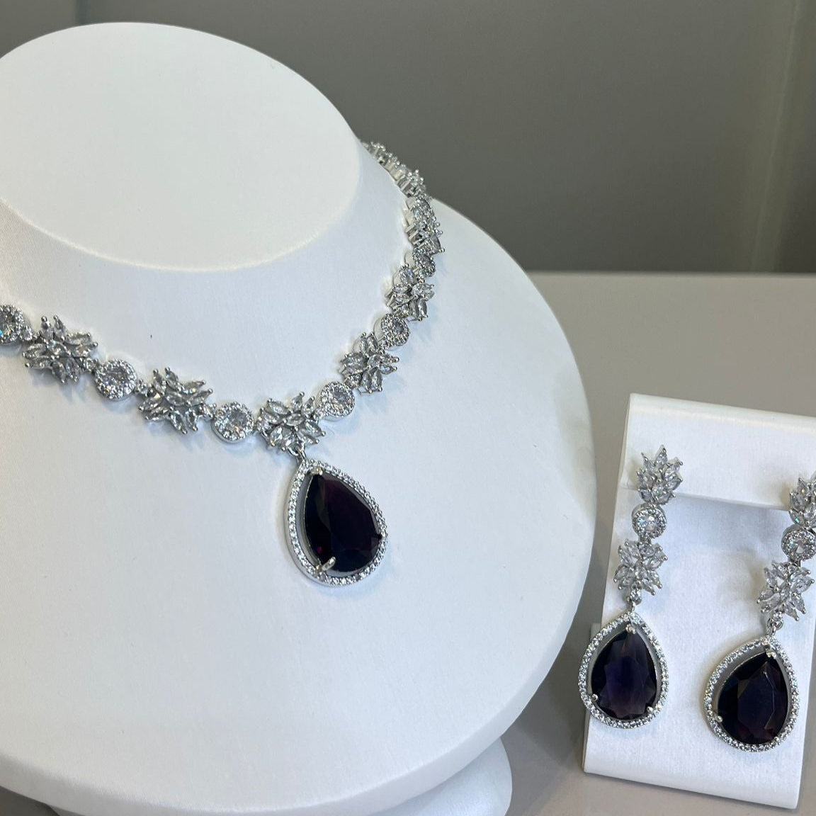 A silver-tone necklace with a large black teardrop gemstone centerpiece and sparkling crystal embellishments, accompanied by a pair of earrings that mirror the necklace's design, displayed on a white bust.
