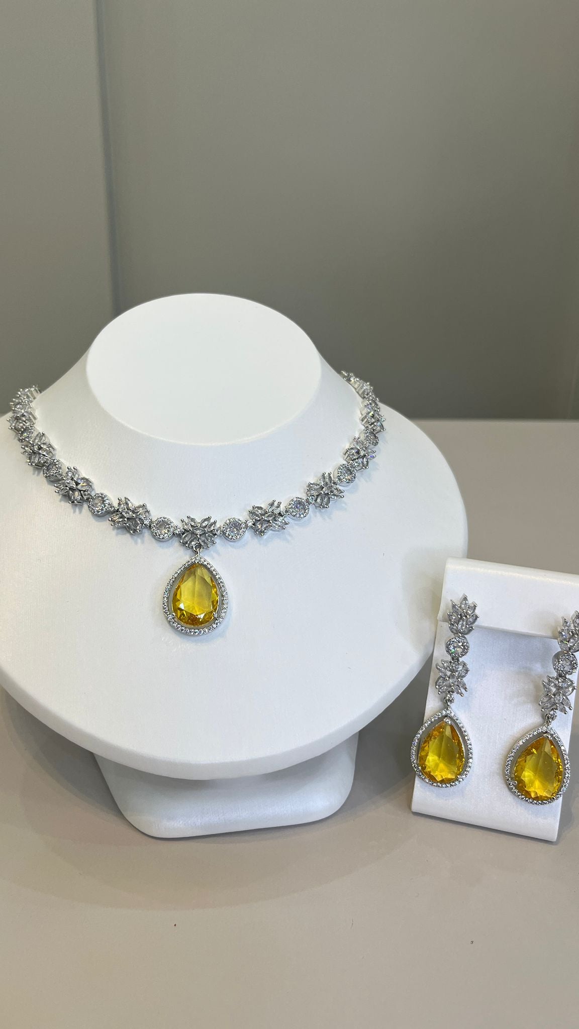 A silver-tone necklace with a large yellow teardrop gemstone centerpiece and sparkling crystal embellishments, accompanied by a pair of earrings that mirror the necklace's design, displayed on a white bust.