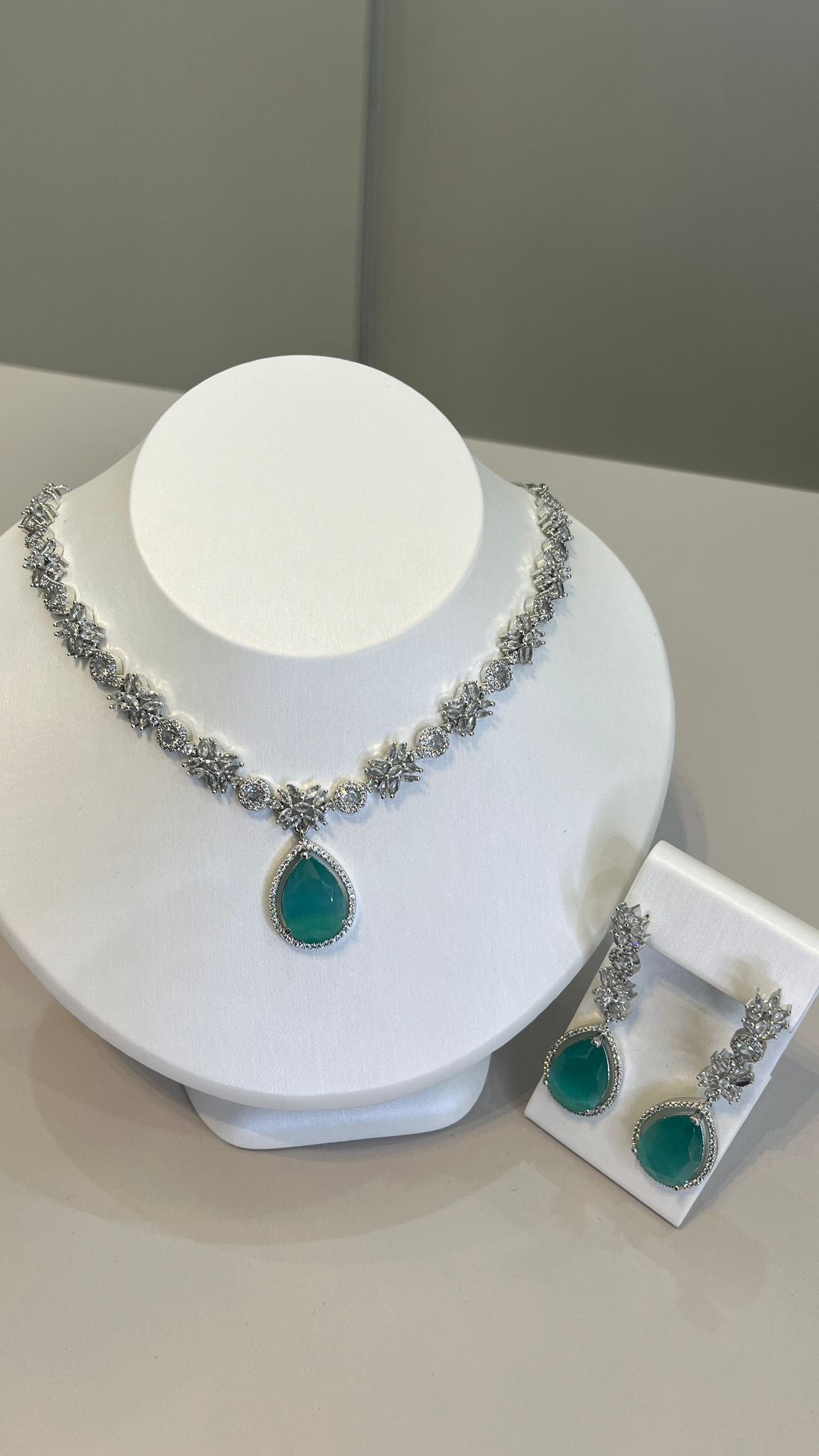 A silver-tone necklace with a large dive-blue teardrop gemstone centerpiece and sparkling crystal embellishments, accompanied by a pair of earrings that mirror the necklace's design, displayed on a white bust.