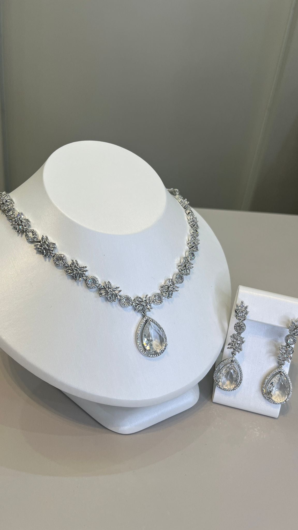 A silver-tone necklace with a large white teardrop gemstone centerpiece and sparkling crystal embellishments, accompanied by a pair of earrings that mirror the necklace's design, displayed on a white bust.