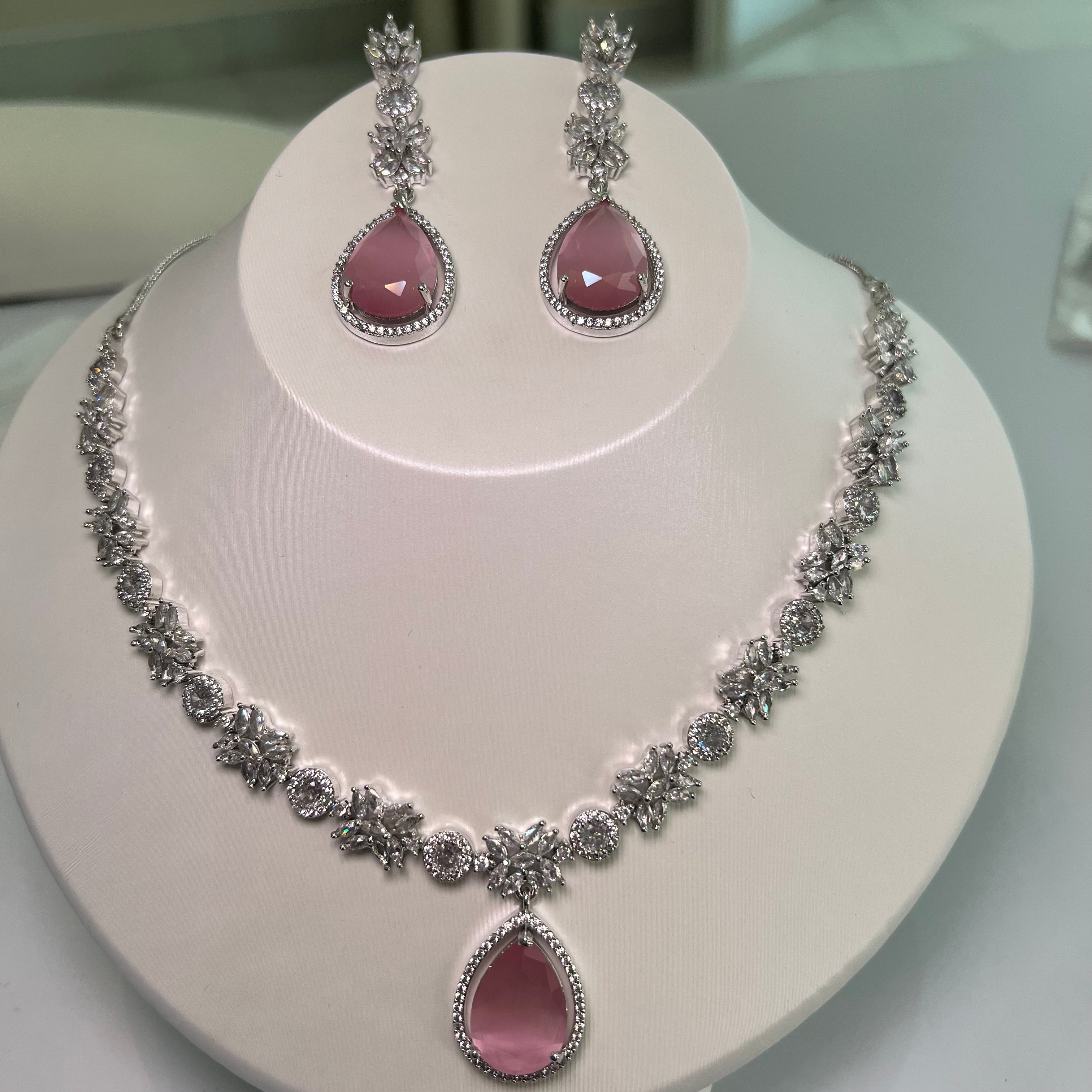 A silver-tone necklace with a large pink teardrop gemstone centerpiece and sparkling crystal embellishments, accompanied by a pair of earrings that mirror the necklace's design, displayed on a white bust.