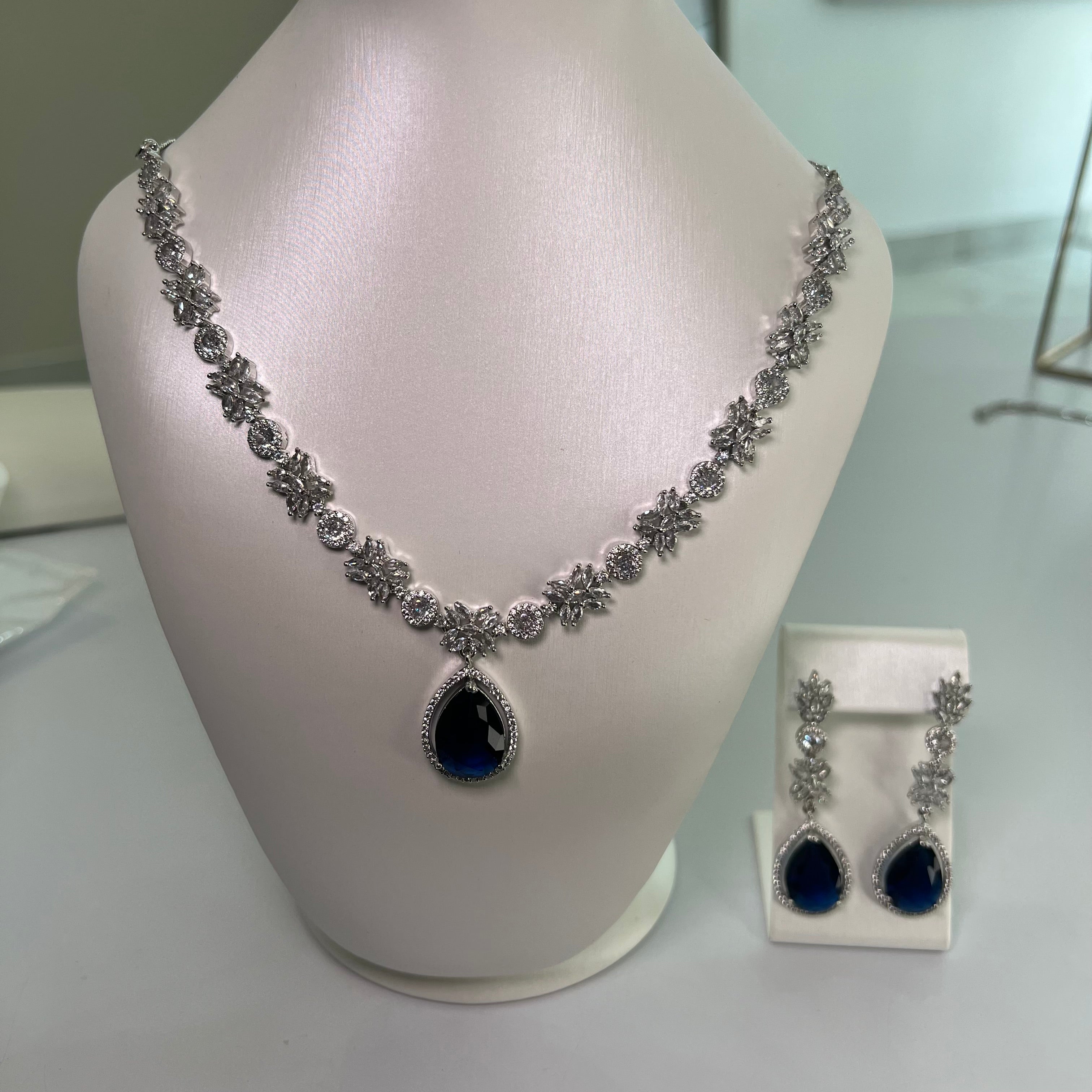 A silver-tone necklace with a large blue teardrop gemstone centerpiece and sparkling crystal embellishments, accompanied by a pair of earrings that mirror the necklace's design, displayed on a white bust.