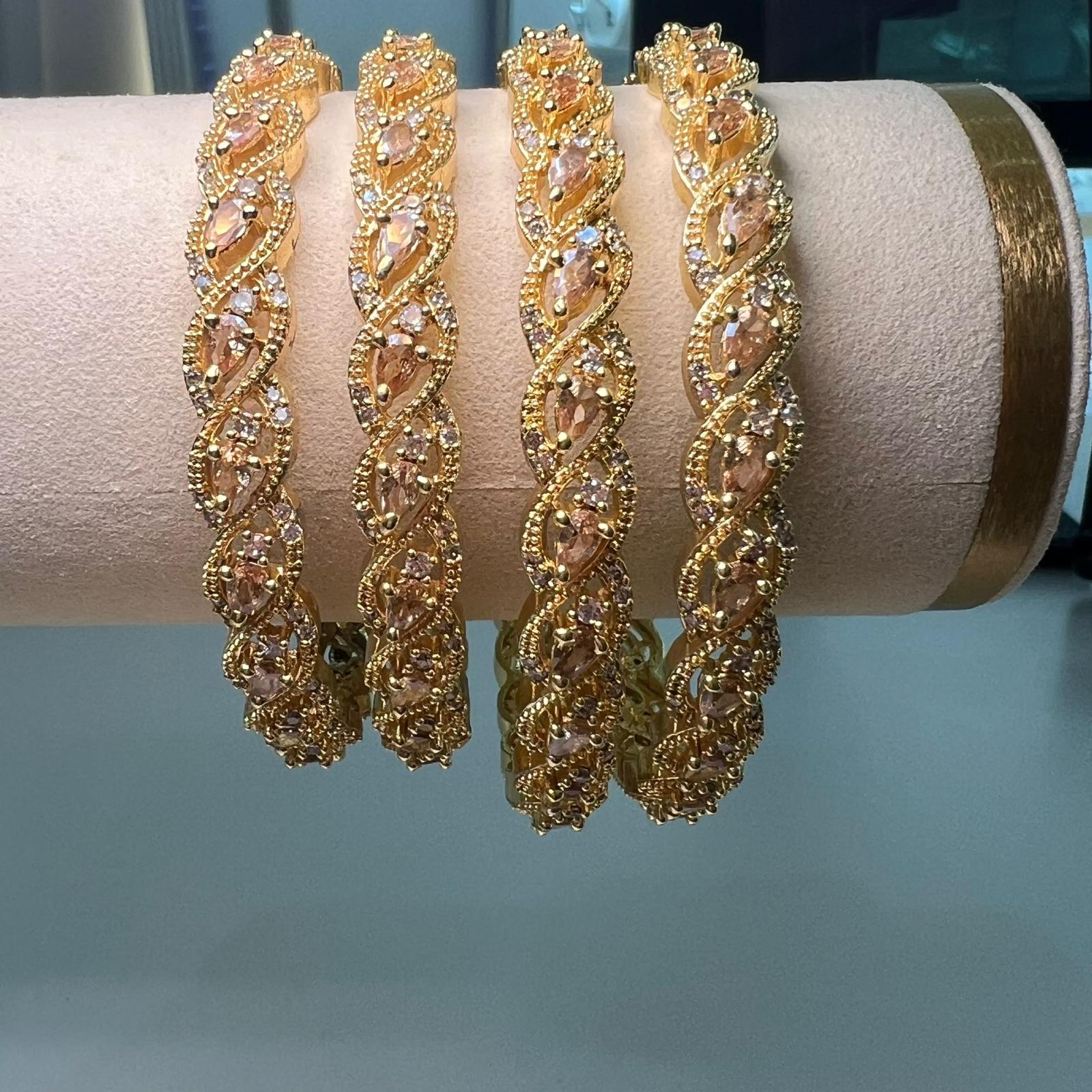 Four gold bracelets with intricate designs on a pink cushion against a light blue background