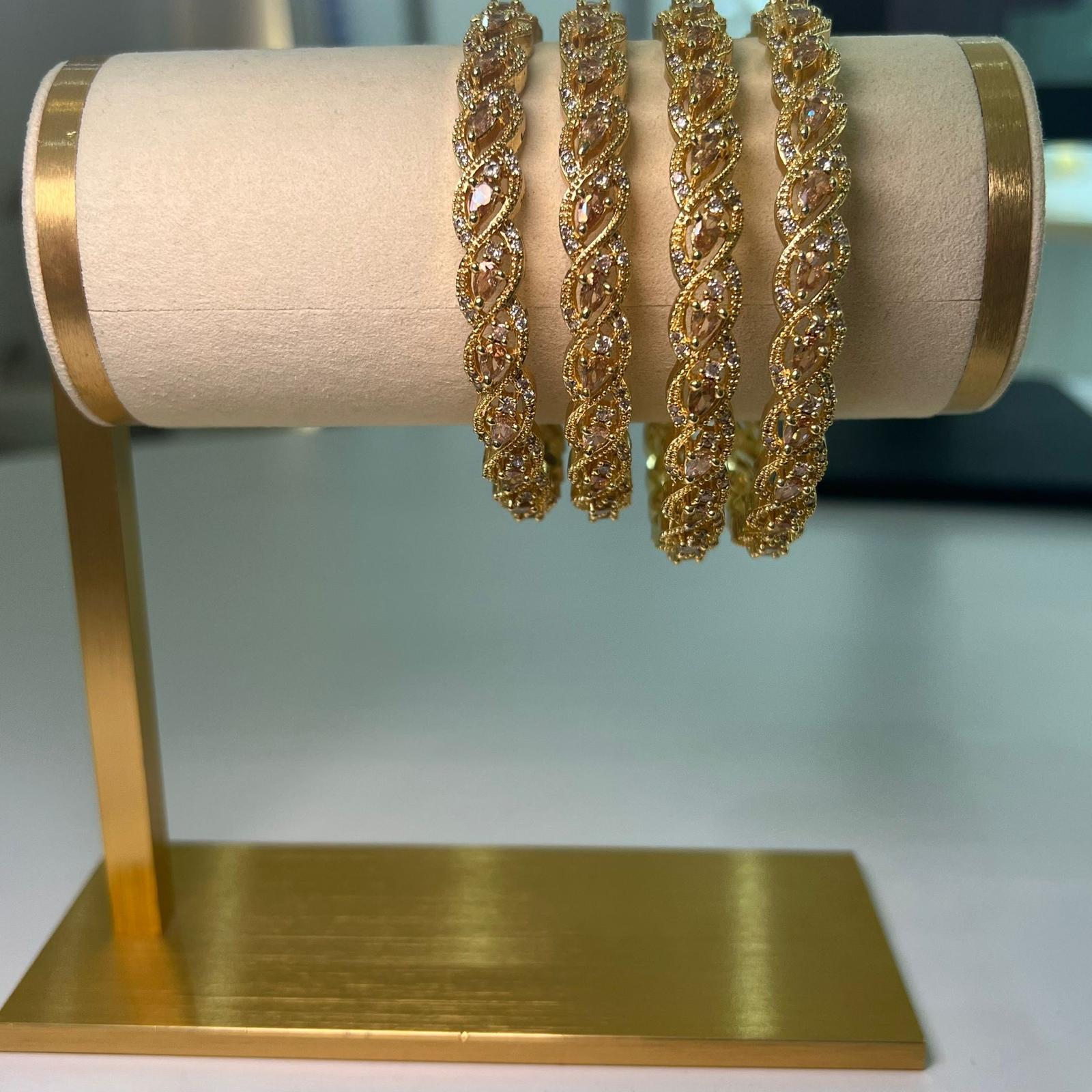 Four gold bracelets with intricate designs on a pink cushion against a light blue background