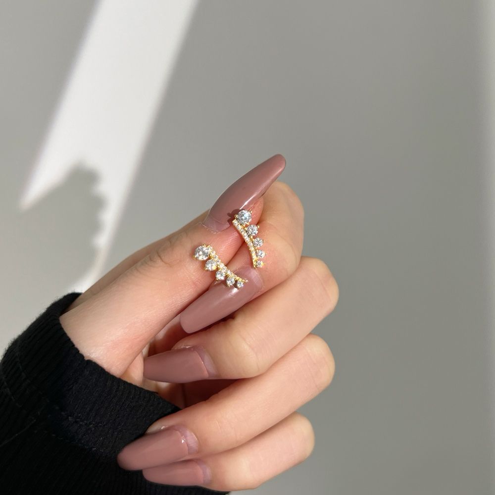 A pair of gold-colored Stone Row earrings featuring multiple round cut clear stones set in a row on a hand with blurred background.