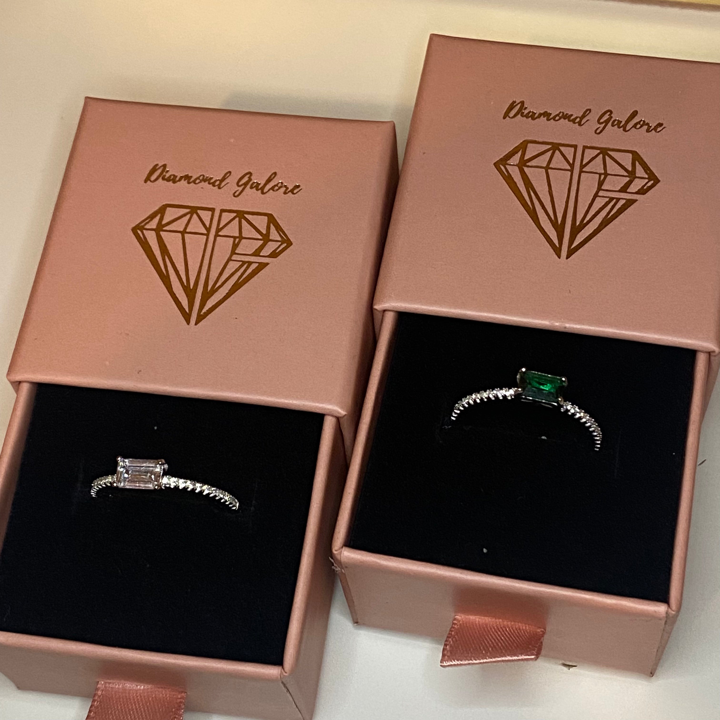 A pair of silver rings featuring a rectangular emerald cut main stone with a surrounding diamond-accented band.