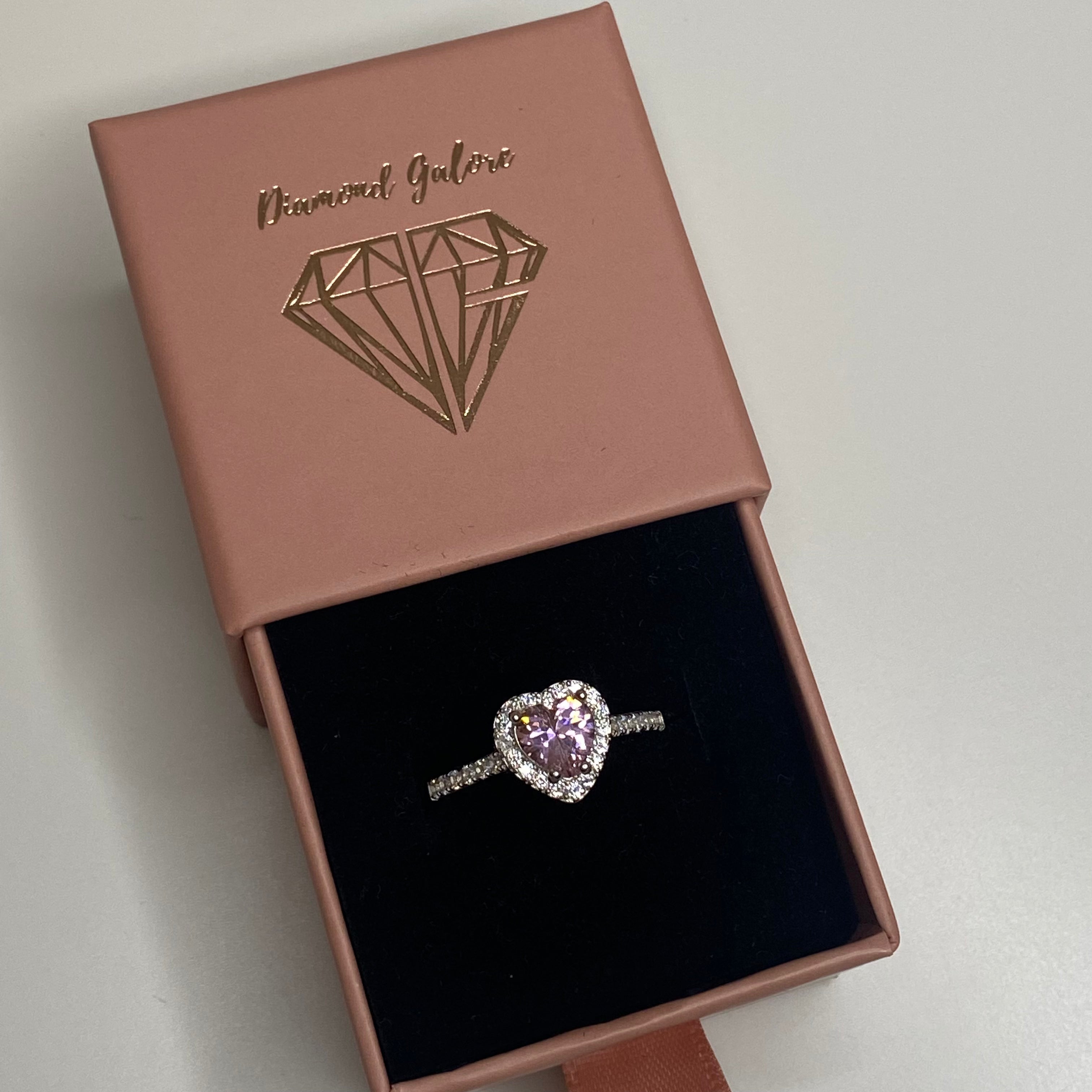 A heart-shaped pink cubic zirconia ring with a halo of smaller clear crystals on a silver band in a ring box.