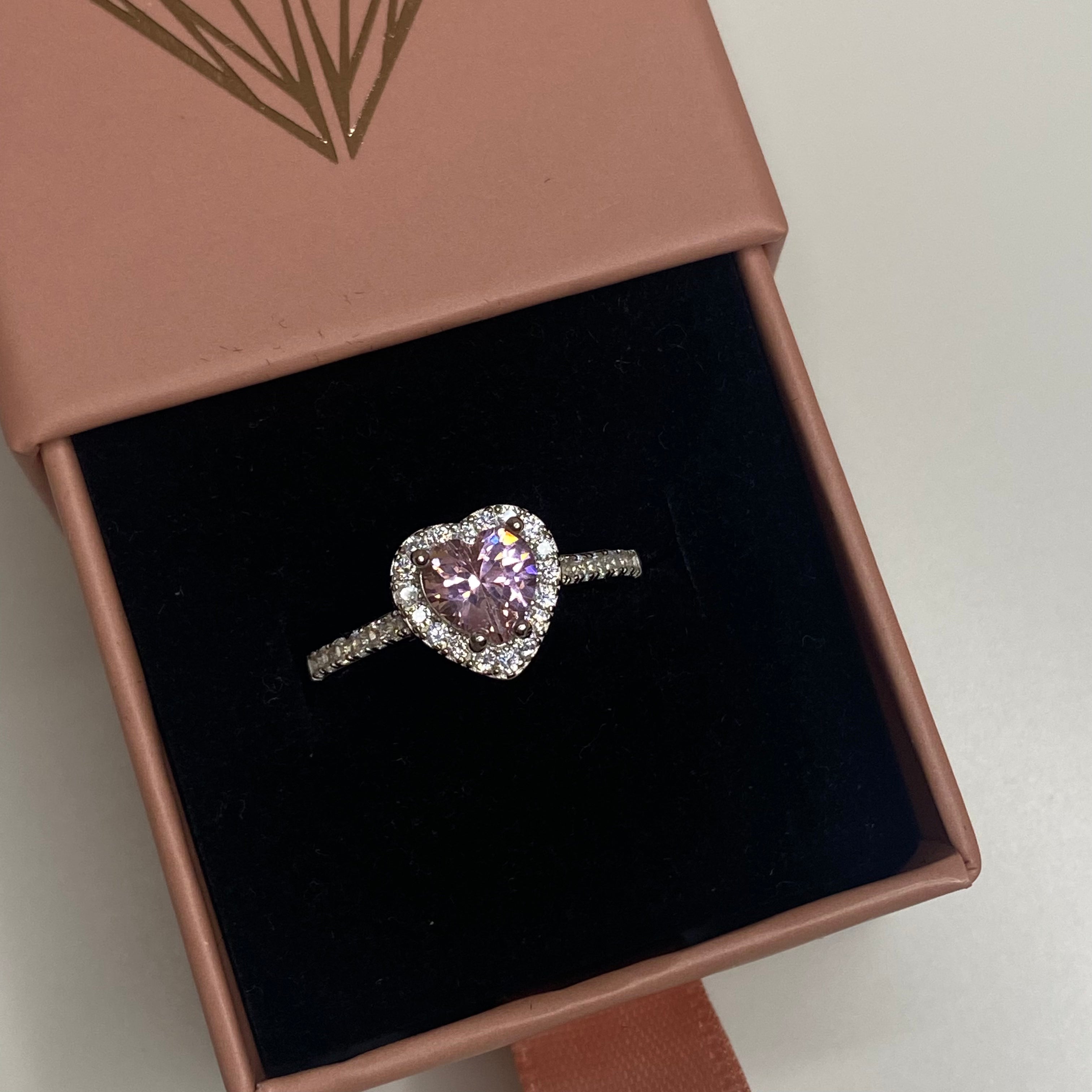 A heart-shaped pink cubic zirconia ring with a halo of smaller clear crystals on a silver band displayed in a box.