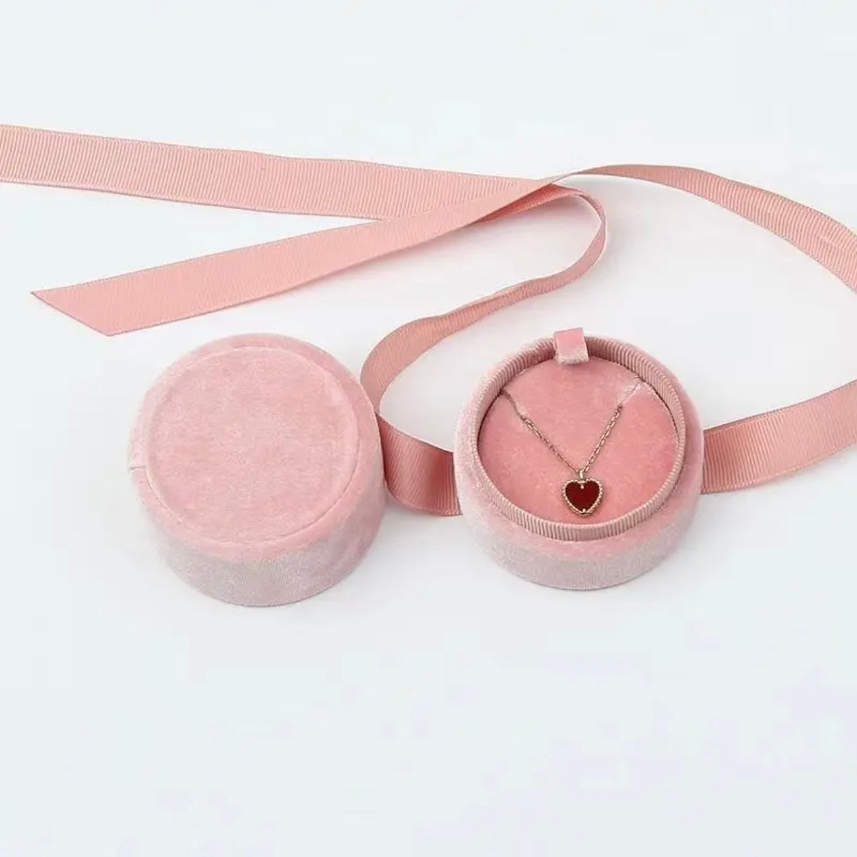 A pink velvet gift box with a ribbon and a red heart necklace inside.
